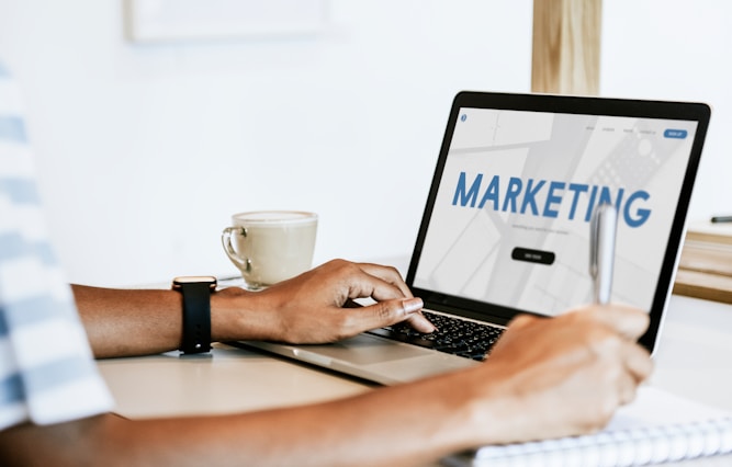 Người làm việc với laptop hiển thị chữ Marketing bên tách cà phê